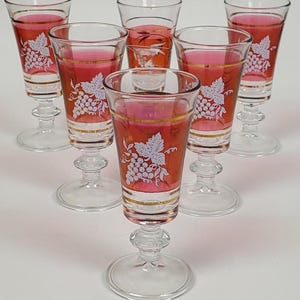 Peut inclure: Ensemble de six verres à vin vintage. Chaque verre a un pied et une base transparents, avec un bol rouge et doré. Un motif de grappe de raisin blanc décore le bol rouge. Les verres sont remplis d'un liquide rose.
