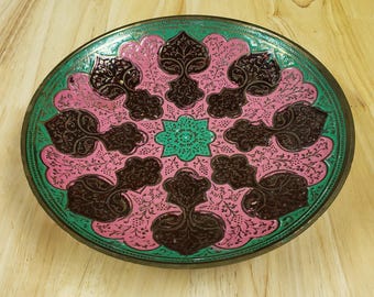 May include: A round, decorative metal plate with an intricate floral design. The plate features a pink center with dark brown and green accents. The outer rim is green, and the plate has a gold-colored edge.