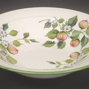 Puede incluir: Un cuenco de cerámica blanca con borde verde. El cuenco está decorado con un motivo floral de frutos rosas y verdes, flores blancas y azules y hojas verdes. El cuenco es redondo y poco profundo.