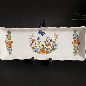 Peut inclure: Un plat de service rectangulaire en céramique blanche avec un bord festonné. Le plat est décoré d'un motif floral et de papillons dans les tons de bleu, orange, jaune et vert. Le plat est soutenu par un support noir.