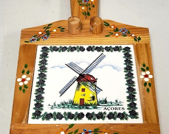 May include: Wooden cutting board with a painted tile insert featuring a yellow windmill with red roof and the word "AÇORES". The wooden frame is decorated with floral and leaf designs. Includes two small wooden pegs and a handle.