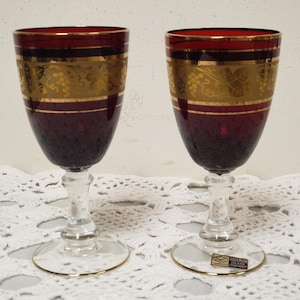 May include: Two ruby red wine glasses with gold trim and a gold grape design. The glasses have clear stems and bases with gold rims. The glasses are on a white lace tablecloth. The glasses have a Bohemian label.