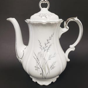 May include: White porcelain teapot with a lid, handle, and spout. The teapot features a floral design in shades of gray and silver trim. The handle and lid handle have a silver finish. The teapot sits on a decorative base.
