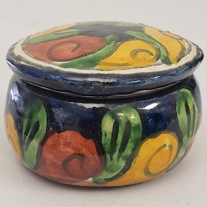 May include: A small, round ceramic trinket box with a lid. The box is painted with a dark blue background and colourful floral designs in yellow, red, and green. The lid has a white border.