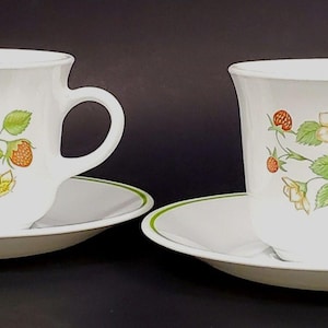 Peut inclure: Deux tasses à thé et soucoupes en céramique blanches. Chaque tasse et soucoupe présente un motif de fraises et de fleurs avec des accents verts et jaunes. Les soucoupes ont une bordure verte. Les tasses ont une anse.
