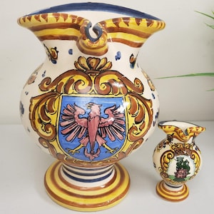 Könnte beinhalten: Zwei Keramikkrüge mit weißem Hintergrund und bunten floralen und geometrischen Mustern. Der größere Krug hat ein blaues und goldenes Wappen mit einem rosa Adler. Der kleinere Krug hat ein grünes und goldenes Wappen mit einem Gebäude.