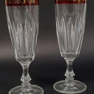 May include: Two clear champagne flutes with a red and gold ornate rim. The flutes have a tall, slender shape with vertical etched lines and sit on a round base. The stem has a decorative design.