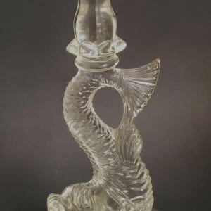 May include: Clear glass candlestick holder with a fish-shaped design. The candlestick has a square base and a detailed fish body with a fin and a circular opening. The top has a floral design to hold a candle.