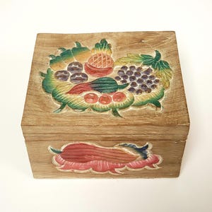 May include: Wooden box with a carved fruit design. The lid features a colourful relief of grapes, a pineapple, and other fruits. The box is rectangular and has a light brown finish. The front of the box has a carved floral design.