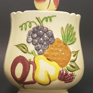May include: A ceramic cookie jar with a fruit design. The jar is cream coloured with a brown, yellow, green, purple, and red fruit design. The lid is a single fruit with a red and yellow design.