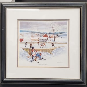 Puede incluir: Impresión enmarcada de una escena invernal con un partido de hockey. La obra de arte representa a niños jugando al hockey sobre una pista al aire libre, con un paisaje nevado y edificios al fondo. El texto "Where Legends Begin" es visible.