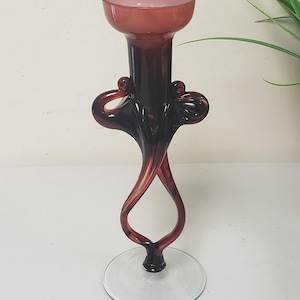 Könnte beinhalten: Ein eleganter Kerzenhalter aus Glas. Der obere Teil ist zartrosa, der Stiel tief burgunderfarben mit wirbelnden Details. Der Sockel ist eine transparente, kreisförmige Scheibe. Das Design ist künstlerisch und dekorativ.