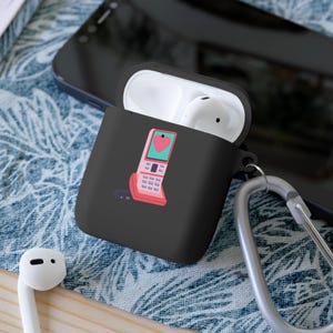 May include: Black phone case with a pink retro phone with a heart on the screen. The case has a white earbud and a silver carabiner.