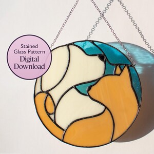 May include: A circular stained glass pattern depicting a dog and cat in cream, orange, and teal. The design is outlined in black and hangs from a silver chain. Text on the left reads "Stained Glass Pattern Digital Download."