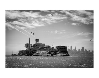 May include: A black and white photograph of Alcatraz Island surrounded by water, featuring the iconic water tower, a tall chimney, and historic prison buildings. Several seagulls fly in the sky above the island, with the San Francisco skyline visible in the distance.