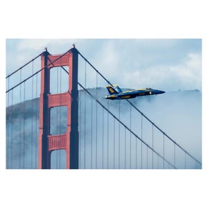 May include: A blue and yellow fighter jet flies over the Golden Gate Bridge in San Francisco, California. The bridge is red and has a large tower in the foreground. The jet is flying through a layer of fog.