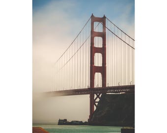 May include: The Golden Gate Bridge in San Francisco, California, is depicted in a scenic photograph. The iconic red bridge is partially obscured by fog, with the blue sky visible above. The bridge spans across the water, with a small island and boats in the distance.