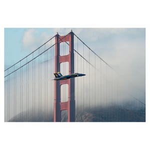 May include: A blue and yellow fighter jet flies over the Golden Gate Bridge in San Francisco, California. The bridge is red and has a white fog rolling in.