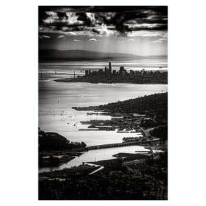 May include: A black and white aerial photograph of a city skyline with a body of water in the foreground. The city is in the distance and the water is filled with boats and ships. The sky is cloudy and the sun is shining through the clouds.