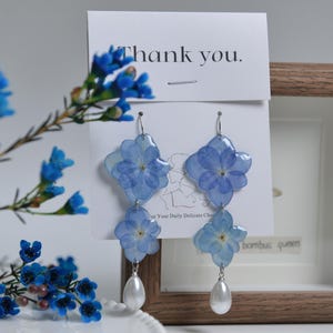 Puede incluir: Un par de pendientes de flores azules con detalles de perlas. Los pendientes se muestran en una tarjeta blanca con el texto "Gracias."