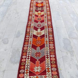 May include: A long, narrow red, orange, and cream colored rug with a geometric pattern. The rug has a fringe on both ends.