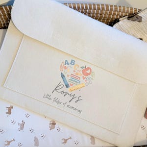 May include: A cream-coloured felt folder with a flap closure. The front displays a heart-shaped design with school-themed illustrations and the name "Rory's" with the text "Little folder of memories". The folder is on patterned fabric.