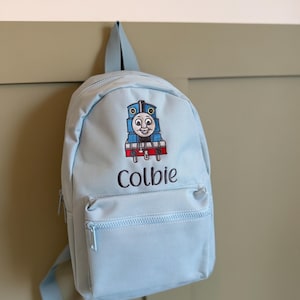 May include: A light blue backpack featuring an embroidered graphic of a blue train engine with a smiling face. Below the train, the name Colbie is embroidered in dark thread. The bag includes a main compartment and a smaller front zippered pocket.