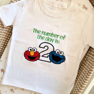 May include: White short-sleeved t-shirt with the text "The number of the day is:" in green. Below the text is a large number "2" with red and blue cartoon character faces on either side.