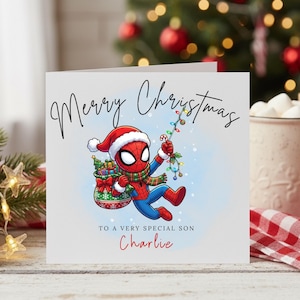 May include: A square white greeting card featuring a cartoon Spider-Man wearing a Santa hat and a green plaid scarf. He carries a sack of gifts and holds a string of colorful Christmas lights. Text reads Merry Christmas, TO A VERY SPECIAL SON, Charlie.