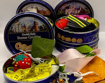 May include: Four blue and gold cookie tins with lids. The tins are filled with various sewing supplies, including buttons, ribbon, thread, and a pincushion. The tins are labeled "Cambridge & Thames" and "Danish Style Butter Cookies".