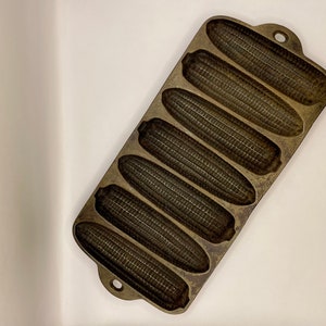 May include: A vintage cast iron cornbread pan with eight individual molds shaped like ears of corn. The pan is brown and has a rustic look.