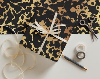 May include: A wrapped gift box with black and gold abstract pattern, tied with a cream ribbon. Scissors, tape, and a pen are arranged nearby. The wrapping paper has a gold and black design.