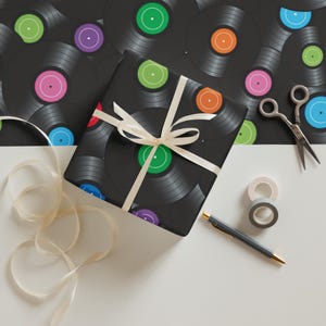 May include: Black wrapping paper with a vinyl record design, featuring colorful center labels. A wrapped gift with a cream ribbon sits on a white surface, along with scissors, tape, and a pen. The background shows more vinyl records.