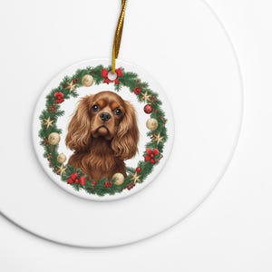 May include: A round, white ceramic Christmas ornament featuring a Cavalier King Charles Spaniel dog surrounded by a festive wreath. The wreath includes green foliage, red bows, gold stars, and ornaments. A gold cord is attached for hanging.