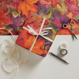 May include: A wrapped gift box with a fall leaf pattern, tied with a cream-colored ribbon. The background is filled with colorful autumn leaves. A pair of scissors, tape, and a pen are also present, suggesting gift-wrapping supplies.