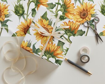May include: A wrapped gift with a cream ribbon, scissors, tape, and a pen on a white surface. The wrapping paper features a watercolor pattern of yellow sunflowers and green leaves on a white background. The ribbon is beige.