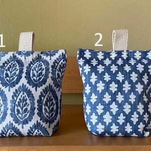 May include: Two fabric doorstops with a loop handle. One doorstop has a blue and white paisley pattern, and the other has a blue and white floral pattern. The doorstops are sitting on a wooden surface.