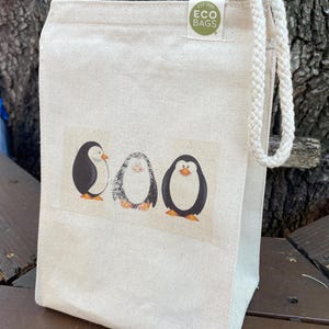 May include: A white canvas tote bag with a drawstring closure. The bag has a green circle logo that says "EST 1965 ECO BAGS". The bag features a graphic of three penguins.