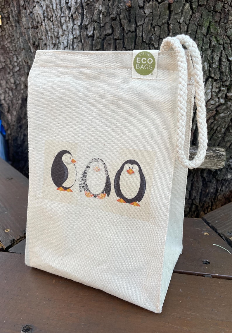 May include: A white canvas tote bag with a drawstring closure. The bag has a green circle logo that says "EST 1965 ECO BAGS". The bag features a graphic of three penguins.