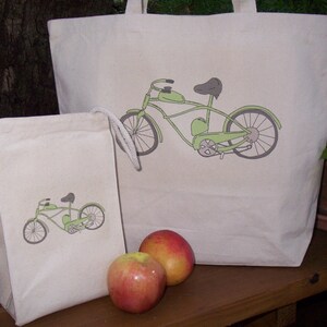 May include: Two canvas tote bags with green bicycle designs. The larger bag has a bicycle with a single seat, while the smaller bag has a bicycle with a double seat. Both bags have white drawstrings. Two red apples are in front of the bags.