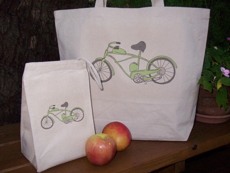 May include: Two canvas tote bags with green bicycle designs. The larger bag has a bicycle with a single seat, while the smaller bag has a bicycle with a double seat. Both bags have white drawstrings. Two red apples are in front of the bags.