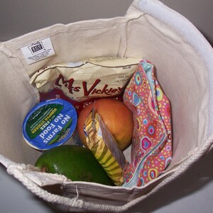 Puede incluir: Una bolsa de lona blanca con una etiqueta marr&oacute;n que dice "Eco Bag" est&aacute; llena de comestibles. Los comestibles incluyen un recipiente de yogur, una bolsa de patatas fritas, un melocot&oacute;n y una bolsa de tela colorida.
