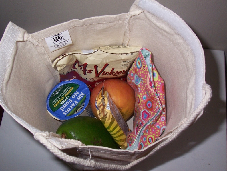 Puede incluir: Una bolsa de lona blanca con una etiqueta marr&oacute;n que dice "Eco Bag" est&aacute; llena de comestibles. Los comestibles incluyen un recipiente de yogur, una bolsa de patatas fritas, un melocot&oacute;n y una bolsa de tela colorida.