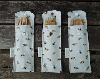 May include: Three beige fabric utensil cases with a repeating pattern of yellow and black bees. Each case contains wooden forks, spoons, and knives. The cases are closed with a snap closure and have a small tag that says "Bag it".