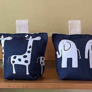 May include: Four doorstop cushions with animal designs. Two are gray with white giraffes, one is navy blue with white giraffes, and one is light blue with white elephants.