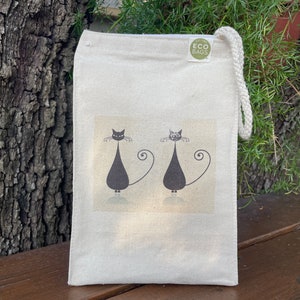 May include: A white canvas tote bag with black cats and the text "Eco Bags" printed on the front. The bag has a drawstring closure.