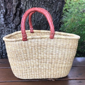 Puede incluir: Una cesta de paja tejida con un acabado natural en color tostado claro y dos asas redondeadas y trenzadas de color marr&oacute;n rojizo. La cesta presenta una parte superior ancha y abierta y una base ovalada estructurada de fibras naturales.