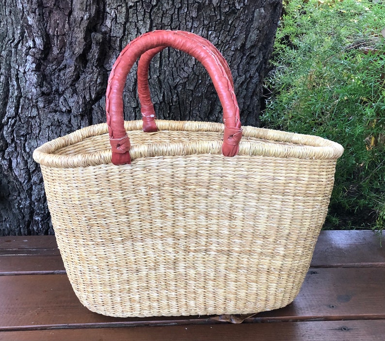 Puede incluir: Una cesta de paja tejida con un acabado natural en color tostado claro y dos asas redondeadas y trenzadas de color marr&oacute;n rojizo. La cesta presenta una parte superior ancha y abierta y una base ovalada estructurada de fibras naturales.