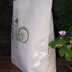 May include: A white canvas tote bag with a green bicycle design printed on the front. The bicycle has a black seat and handlebars.