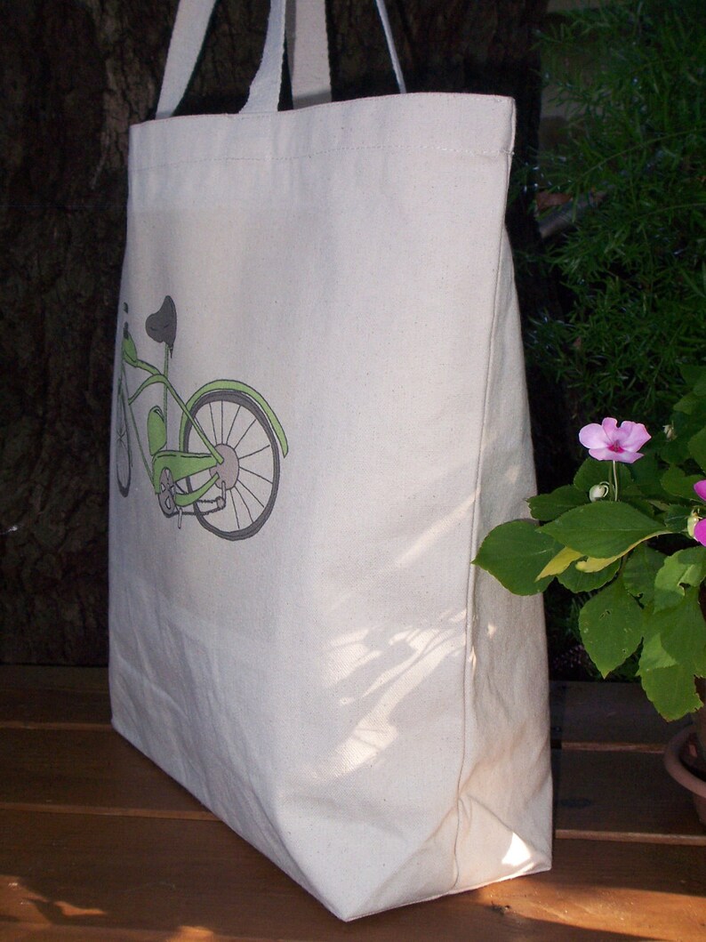 May include: A white canvas tote bag with a green bicycle design printed on the front. The bicycle has a black seat and handlebars.